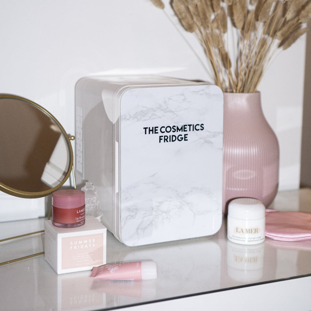 The White Marble Cosmetics Fridge on a vanity table filled with luxury skincare products and sheet mask. Nexy to flowers and makeup, cosmetics and perfume